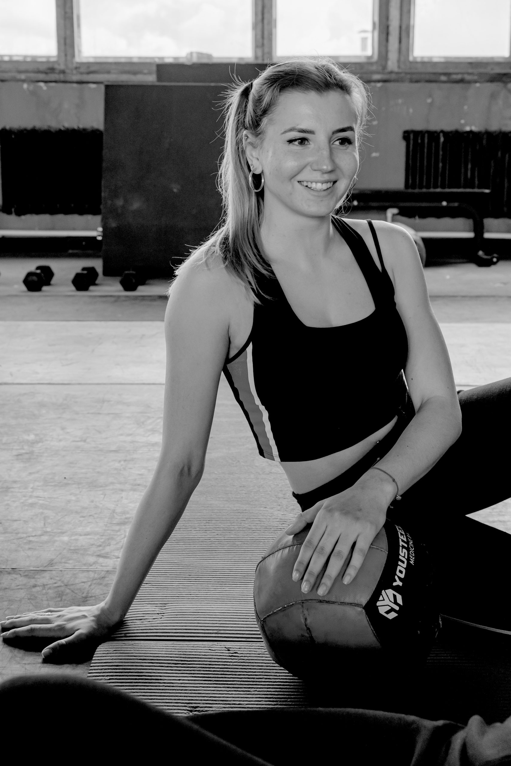 woman sitting in gym to make wall balls easier holding medicine ball and smiling with her hair in a ponytail and gym equipment around her