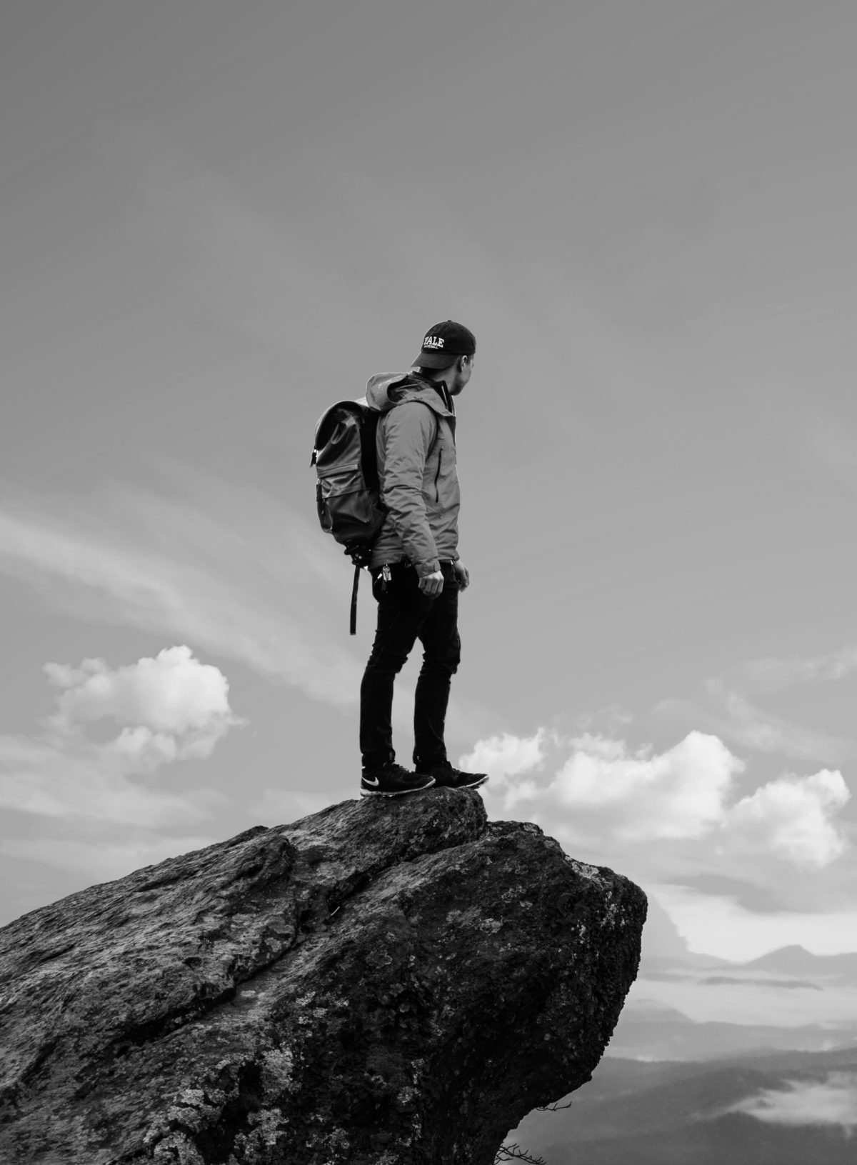 man who can embrace discomfort wearing a backpack and standing on a cliff in the sky looking down at his achievements