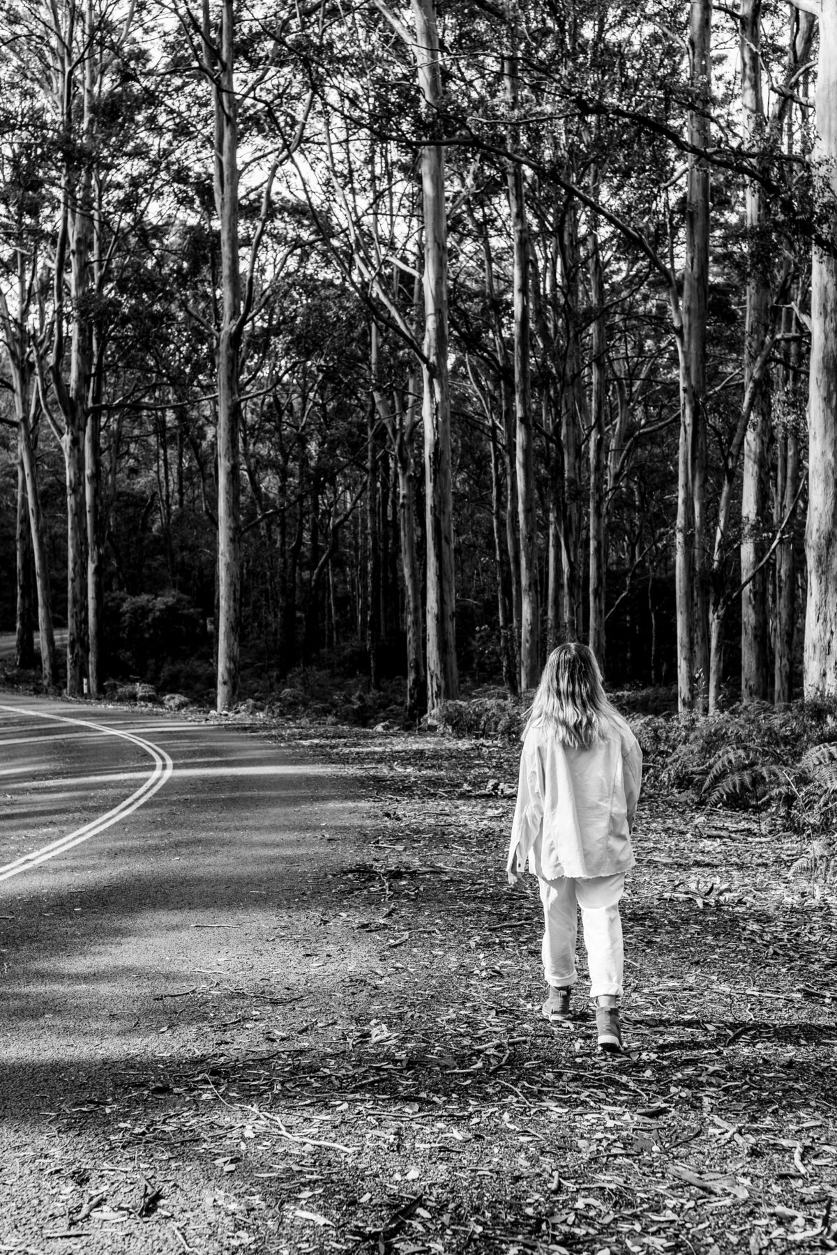 woman wearing white pants and top walking facing away along a road peaceful with mental health benefits of exercise into the trees