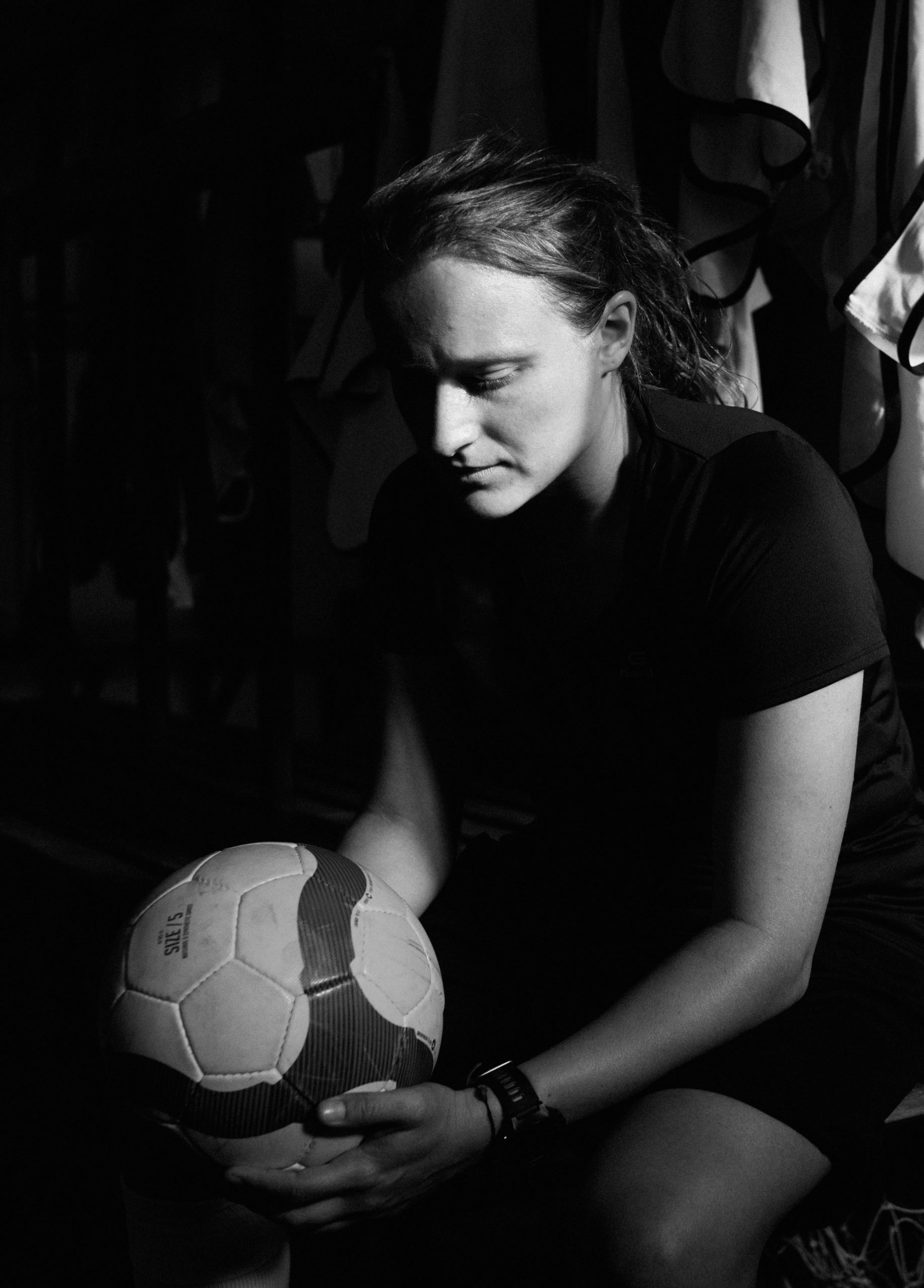 woman with hair in ponytail overcoming feelings of failure sitting in dark room holding soccer ball looking stressed